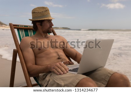Stockfoto: Front View Of Shirtless Young Man Relaxing On Sun Lounger Looking Away While Using Laptop On Beach I
