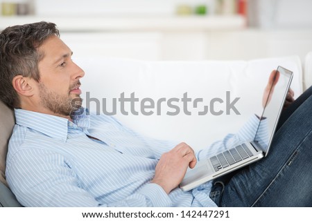 Imagine de stoc: Portrait Of Happy Mature Man Using Laptop Lying On Sofa In House