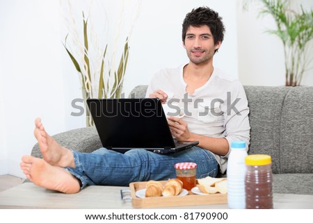 Stock photo: Young Man Seated On A Sofa With Legs Stretched Out Doing Computer And Eating Breakfast