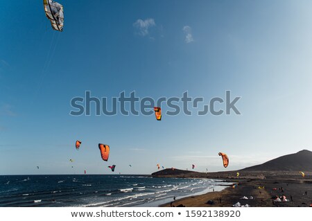 Stockfoto: Many Colorful Kites On Beach And Kite Surfers Riding Waves During Windy Day