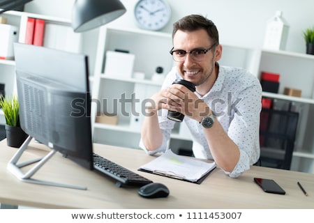 ストックフォト: A Young Man In Glasses Stands Near A Table In The Office Holding A Glass Of Coffee