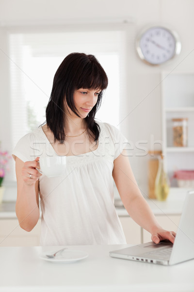 Stock photo: Attractive brunette woman holding a cup of coffee while relaxing with her laptop in the kitchen