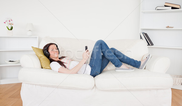Stock photo: Beautiful red-haired female listening to music with headphones while lying on a sofa in the living r