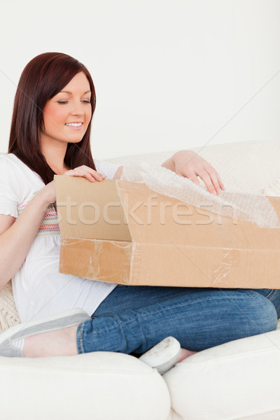 Stock photo: Good looking red-haired woman opening a carboard box while sitting on a sofa in the living room