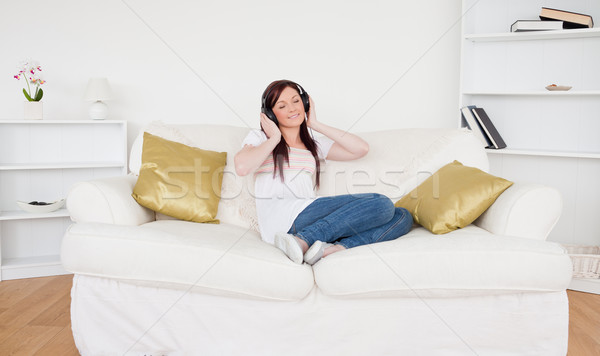 Stock photo: Pretty red-haired female listening to music with headphones while sitting on a sofa in the living ro
