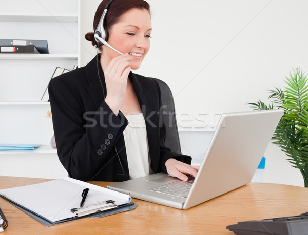 Stock photo: Young pretty red-haired female in suit typing on her laptop and using headphones while sitting in an