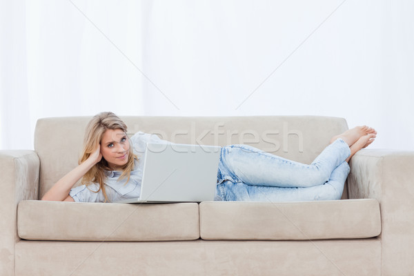 Stock photo: A woman looking at the camera is lying on a couch resting and using her laptop