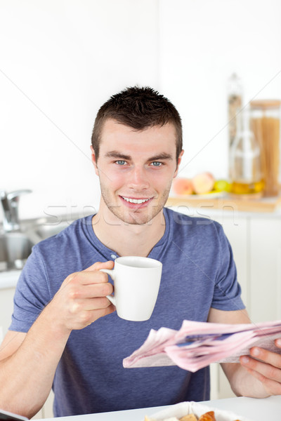 Stock photo: Positive young man holding a cup and a newspaper smiling at the camera sitting in the kitchen