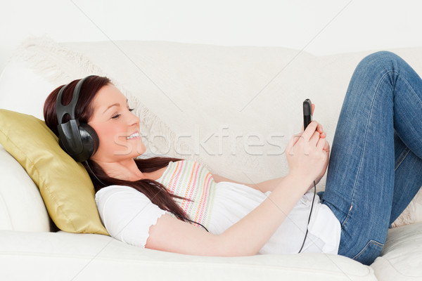 Stock photo: Beautiful red-haired woman listening to music with headphones while lying on a sofa in the living ro