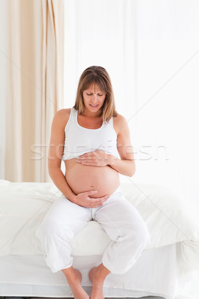 Stock photo: Good looking pregnant female caressing her belly while sitting on a bed in her apartment