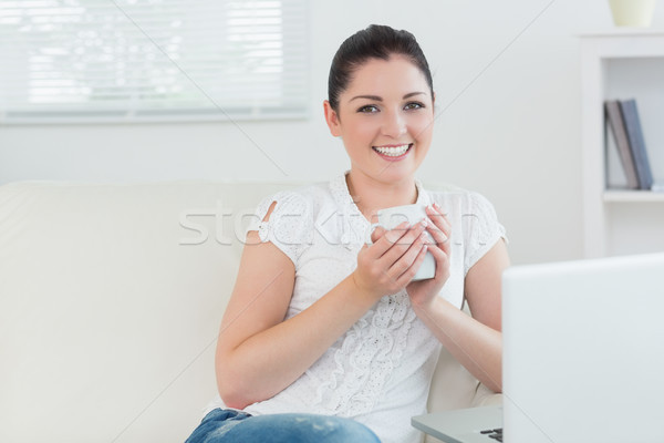 Stock photo: Woman sitting on the couch in a living room and holding a cup while using a laptop