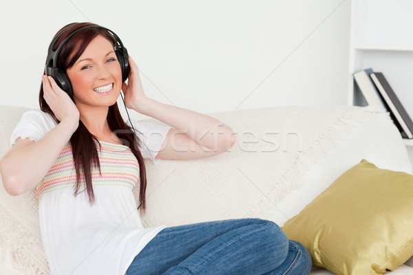 Stock photo: Attractive red-haired woman listening to music with headphones while sitting on a sofa in the living