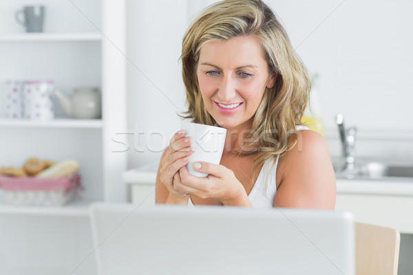 Stock photo: Smiling woman holding mug in kitchen with laptop