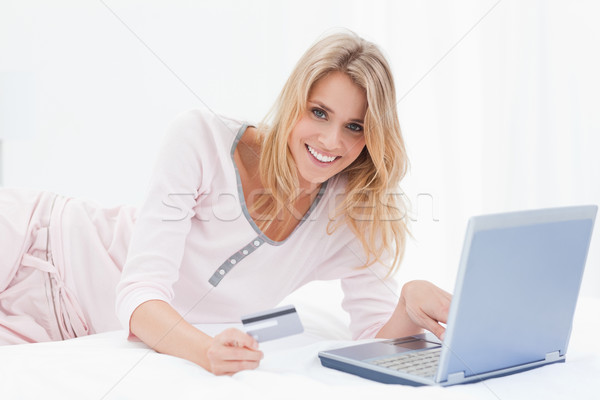 Stock photo: A woman is lying on the bed smiling as she looks forward while ordering items online with her laptop