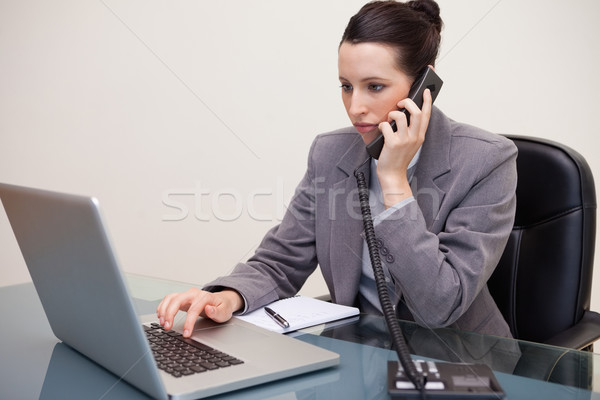 Stock photo: Young businesswoman typing on her laptop while on the telephone