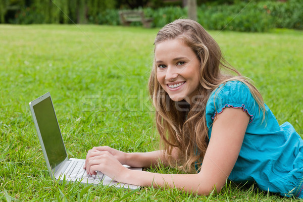 Stock photo: Happy teenager using her laptop while lying in a park and looking at the camera