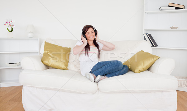 Stock photo: Good looking red-haired female listening to music with headphones while sitting on a sofa in the liv