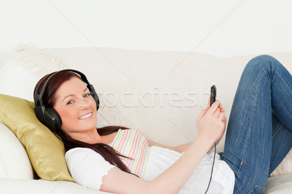 Stock photo: Good looking red-haired woman listening to music with headphones while lying on a sofa in the living