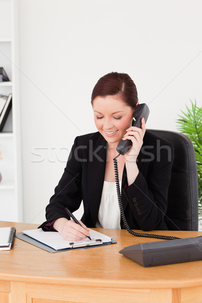 Young attractive red-haired woman in suit writing on a notepad and phoning while sitting in an offic Stock photo © wavebreak_media
