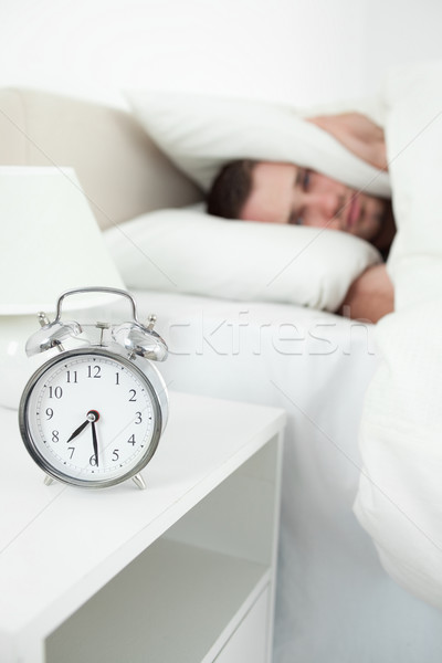 Stock photo: Portrait of a tired young man covering his ears with a pillow while his alarm clock is ringing