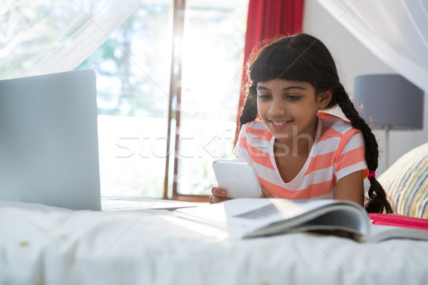Fată telefon mobil carte laptop pat acasă Imagine de stoc © wavebreak_media