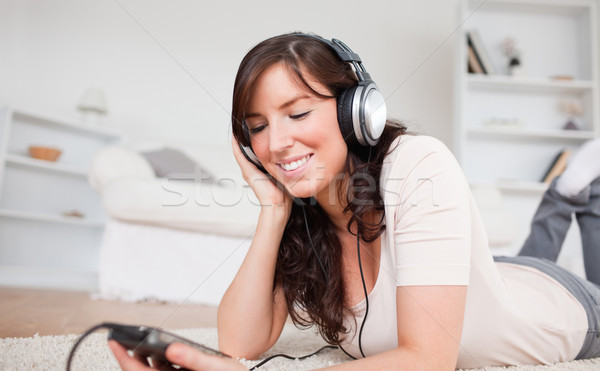 Stock photo: Cute brunette woman listening to music with her mp3 player while lying on a carpet in the living roo