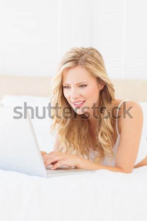 Stock photo: Cheerful young blonde lying on her bed using laptop