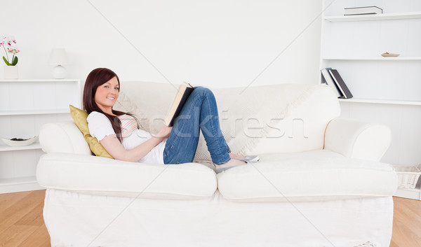 Attractive red-haired woman reading a book while lying on a sofa in the living room Stock photo © wavebreak_media