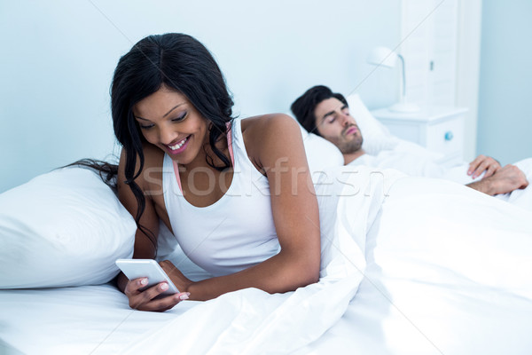 Stock photo: Woman checking her mobile phone while man sleeping on bed