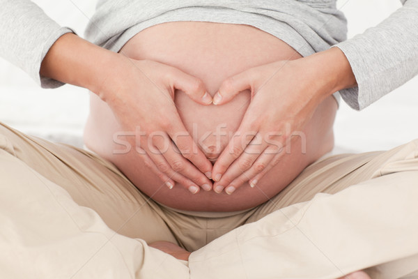 Stock photo: Close up of a pregnant woman doing heart sign on her belly sitting on a bed