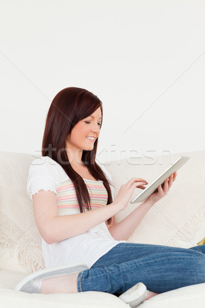 Stock photo: Beautiful red-haired woman relaxing with her tablet while sitting on a sofa in the living room