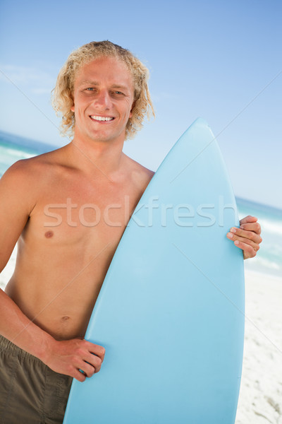 Stock photo: Young smiling man standing upright while holding his perched surfboard