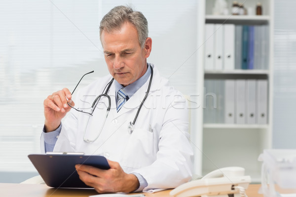 Concentrating doctor sitting at his desk with clipboard Stock photo © wavebreak_media