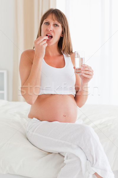 Stock photo: Beautiful pregnant female taking a pill while sitting on a bed in her apartment