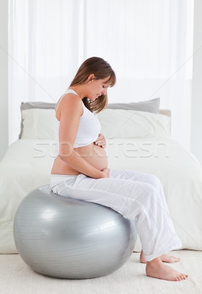 Stock photo: Good looking pregnant female caressing her belly while sitting on a gym ball in her bedroom