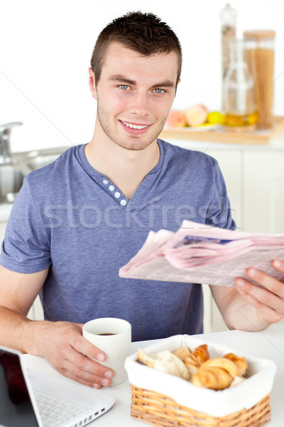 Positive young man holding a cup and a newspaper smiling at the camera sitting in the kitchen at hom Stock photo © wavebreak_media