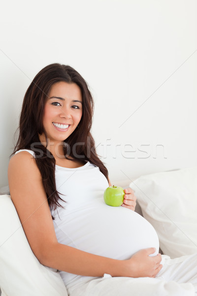 Stock photo: Gorgeous pregnant woman holding an apple on her belly while lying on a bed at home