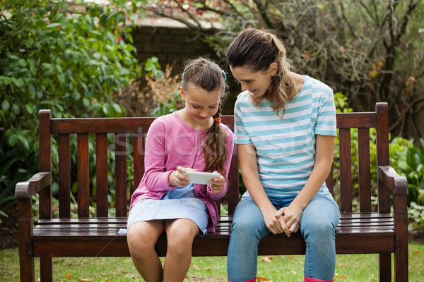 Smiling woman sitting by girl using mobile phone on wooden bench Stock photo © wavebreak_media