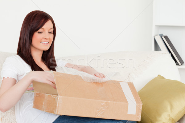 Stock photo: Attractive red-haired woman opening a carboard box while sitting on a sofa in the living room