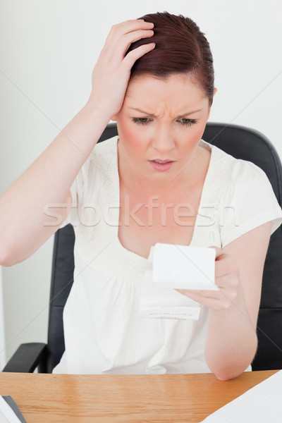 Stock photo: Young attractive red-haired female being stunned by the amount of the receipt while sitting at a des