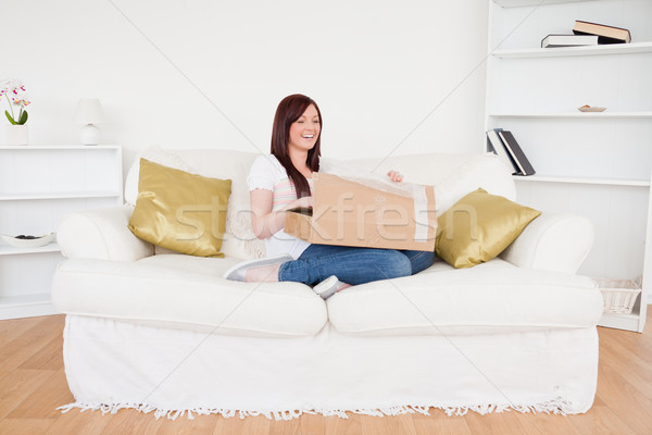 Stock photo: Attractive red-haired female opening a carboard box while sitting on a sofa in the living room
