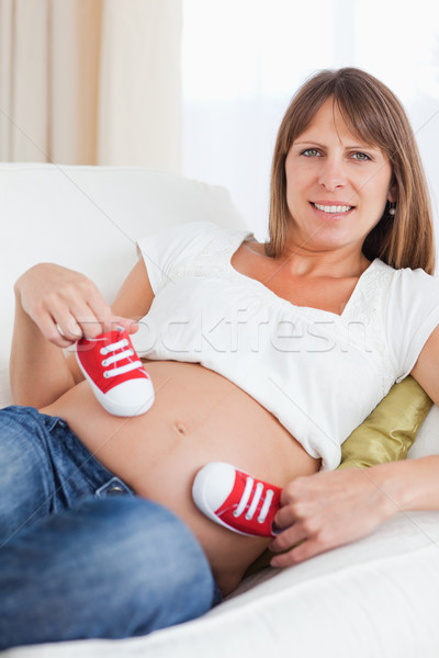 Stock photo: Attractive pregnant female playing with red baby shoes while lying on a sofa in her living room